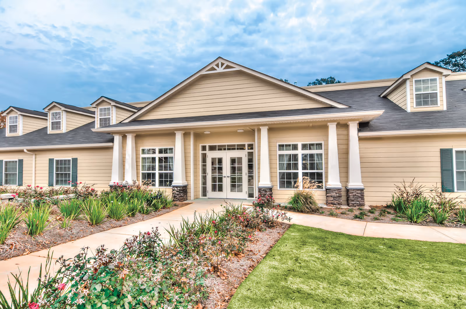Front exterior view of a beige senior living facility building with white columns, multiple windows, and a landscaped garden with flowers and green grass in front under a partly cloudy sky.