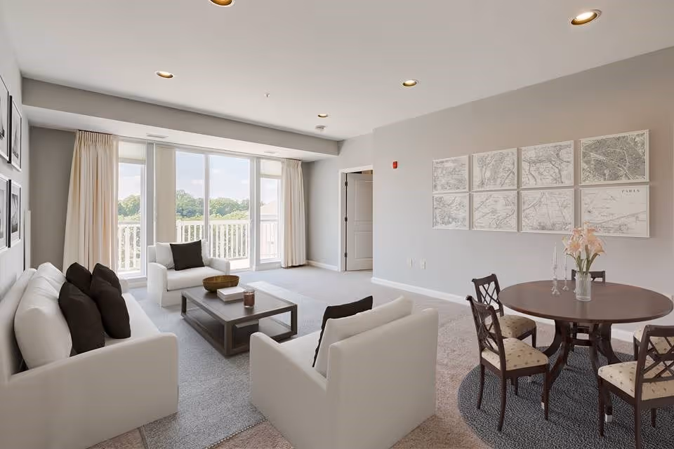 A bright and spacious living room with large windows and sliding glass doors leading to a balcony. The room features white sofas with dark cushions, a wooden coffee table, and a round dining table with four chairs. The walls are light gray and decorated with framed maps and black-and-white photos. The carpet is light gray, and the room is well-lit with recessed ceiling lights.