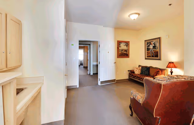 A cozy seating area with upholstered chairs and a sofa, framed artwork and a lamp, with a doorway opening to a hallway.