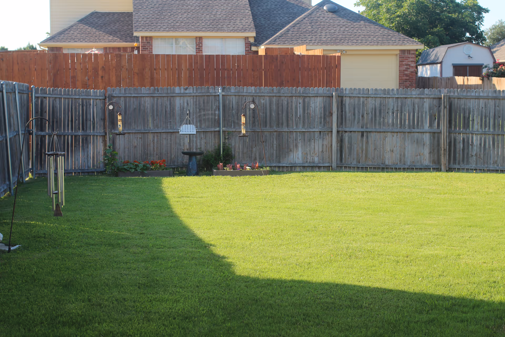 A backyard with a well-maintained green lawn enclosed by a wooden fence. There are bird feeders and a birdbath along the fence, with some small flower beds. Houses and a shed are visible beyond the fence under a clear sky.