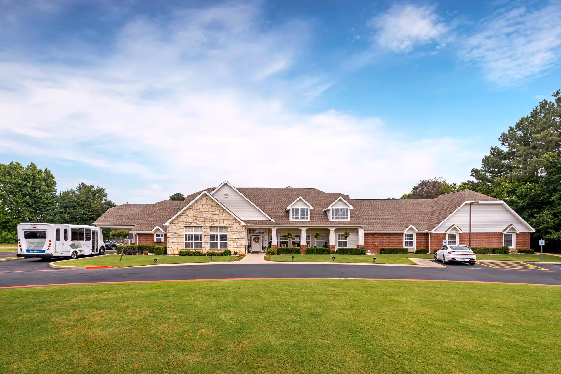 Front exterior view of Brookdale Texarkana facility showing a single-story building with a combination of stone and brick facade, a covered entrance with hanging plants, a parking lot with a white car and a shuttle bus, and a well-maintained green lawn under a partly cloudy blue sky.