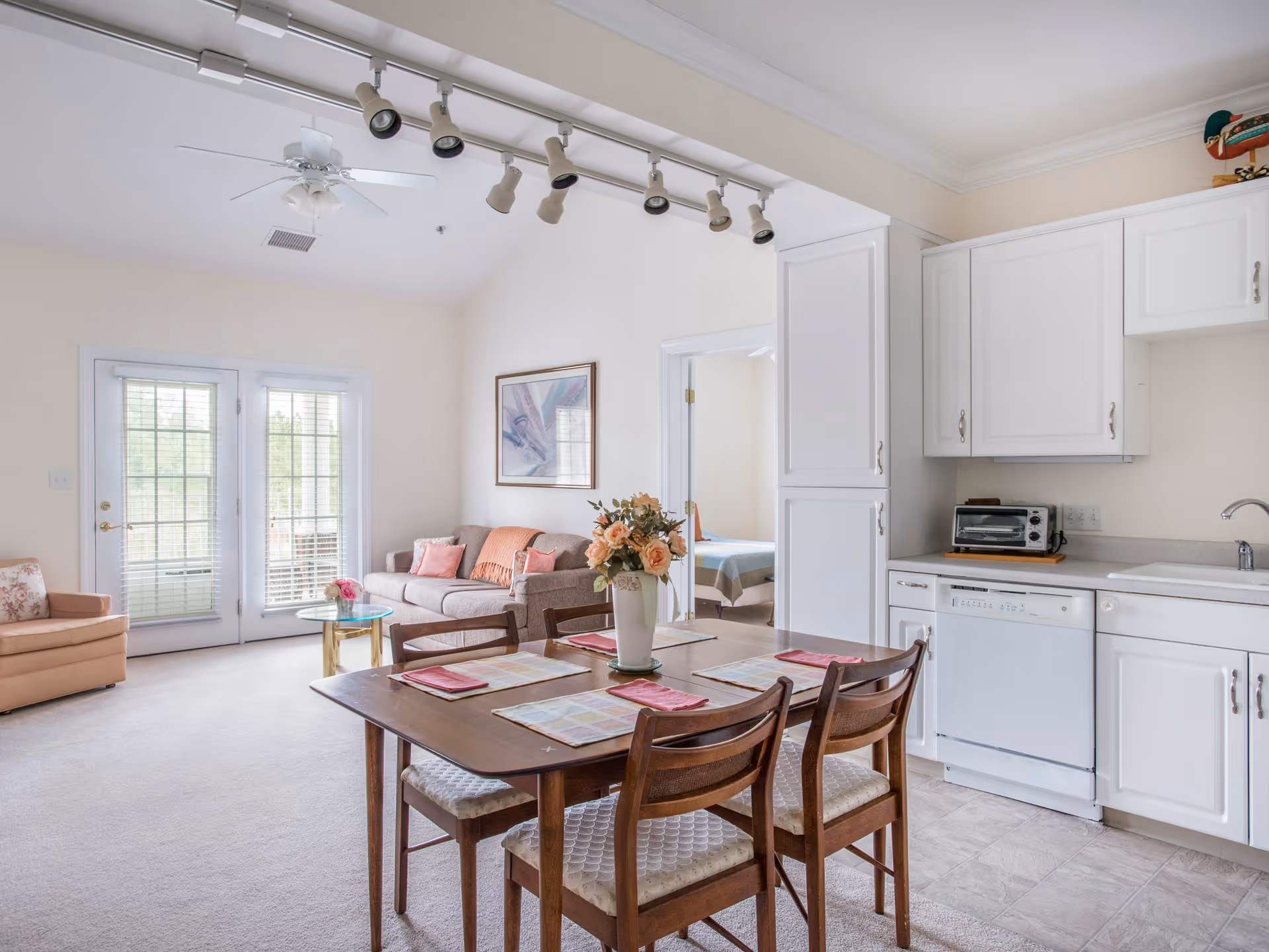 Bright and airy living and dining area with a wooden dining table set for four, a beige sofa with pink cushions, a glass coffee table, an armchair, white kitchen cabinets, a dishwasher, and a toaster oven. French doors with blinds lead outside, and a bedroom is visible through an open doorway.