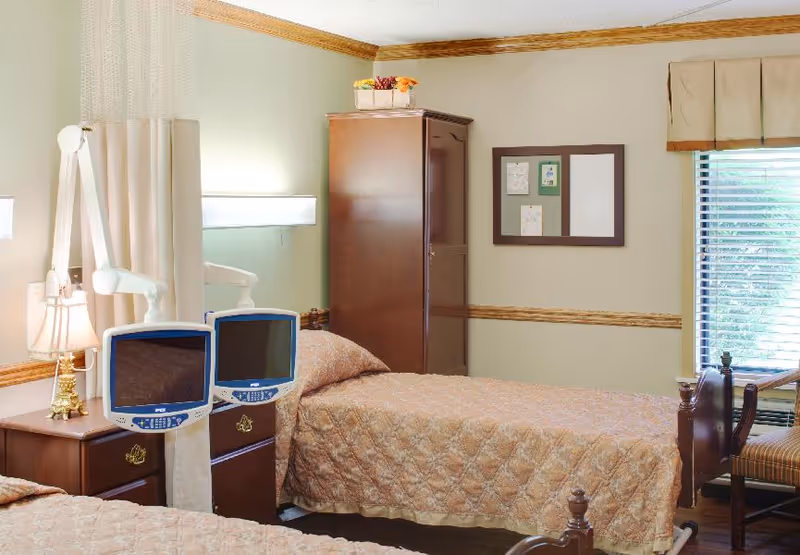 Furnished assisted-living bedroom with two twin beds, bedside monitors, wooden furniture, and a window.