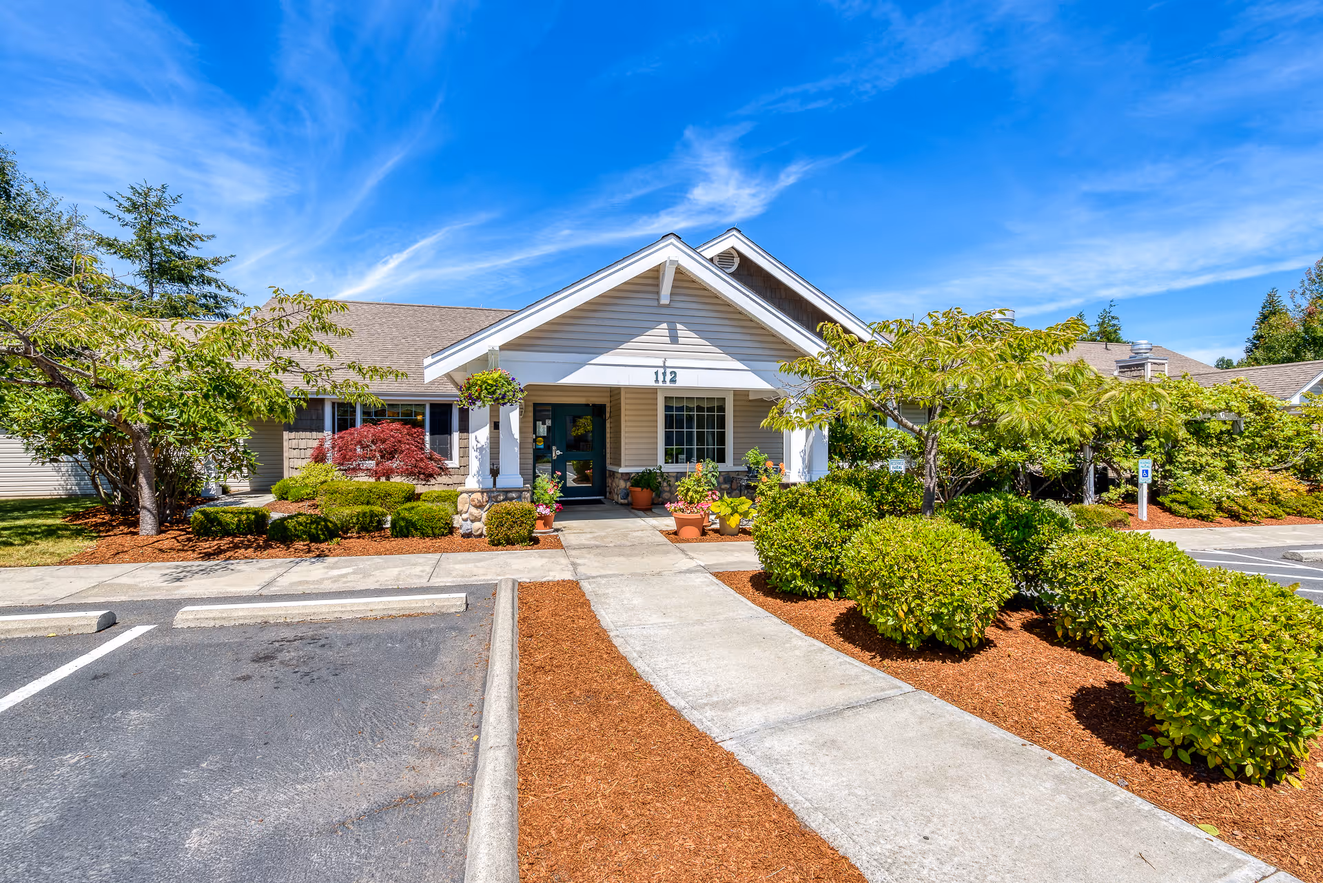 Front exterior view of a single-story building with a gabled roof, surrounded by well-maintained landscaping including bushes, trees, and potted plants. A concrete walkway leads to the entrance under a covered porch. The sky is clear and blue.