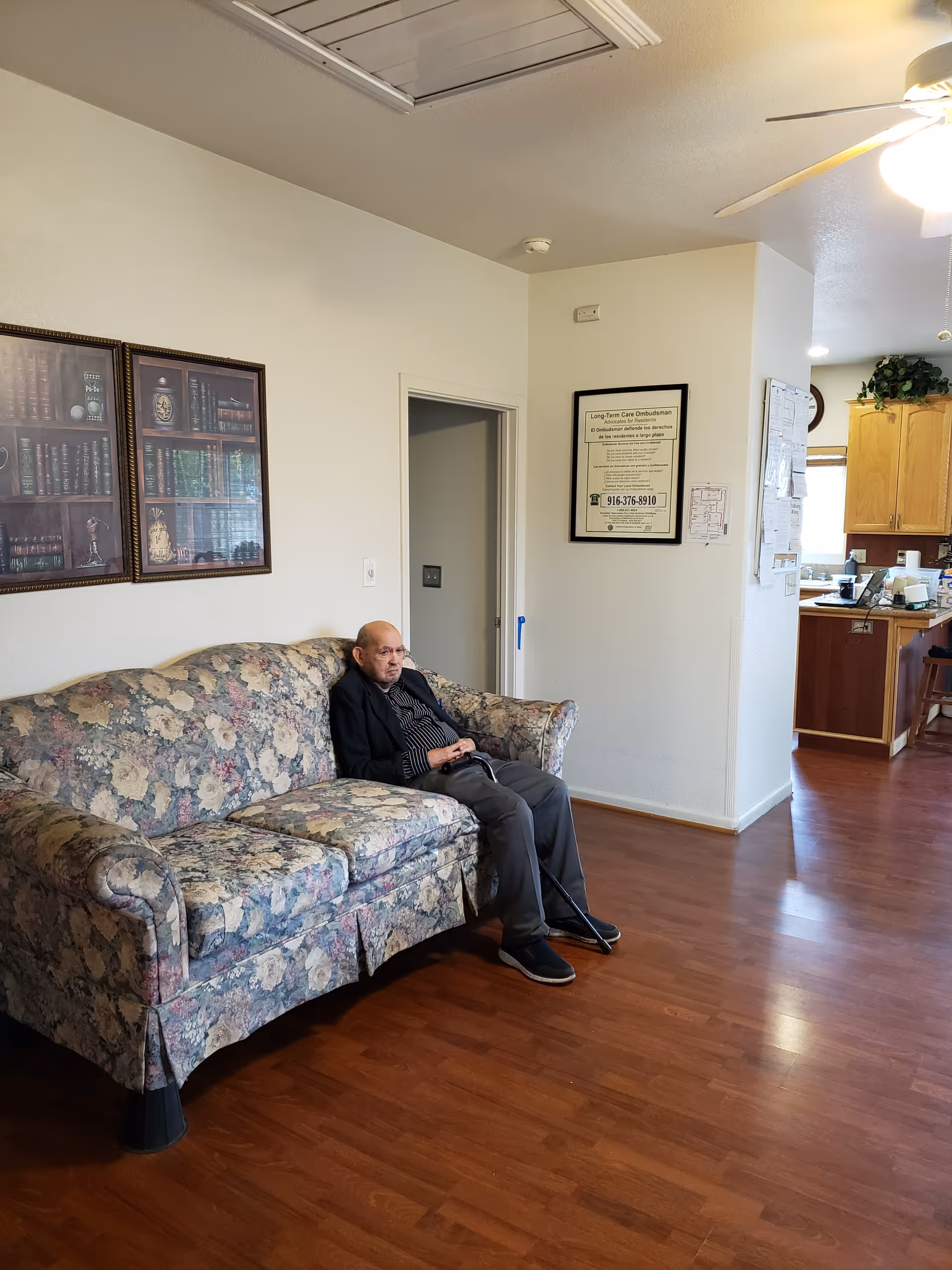 An elderly man sitting on a floral patterned couch in a room with wooden flooring. The room has white walls with framed pictures and a notice on the wall. In the background, there is a kitchen area with wooden cabinets and a ceiling fan with a light.