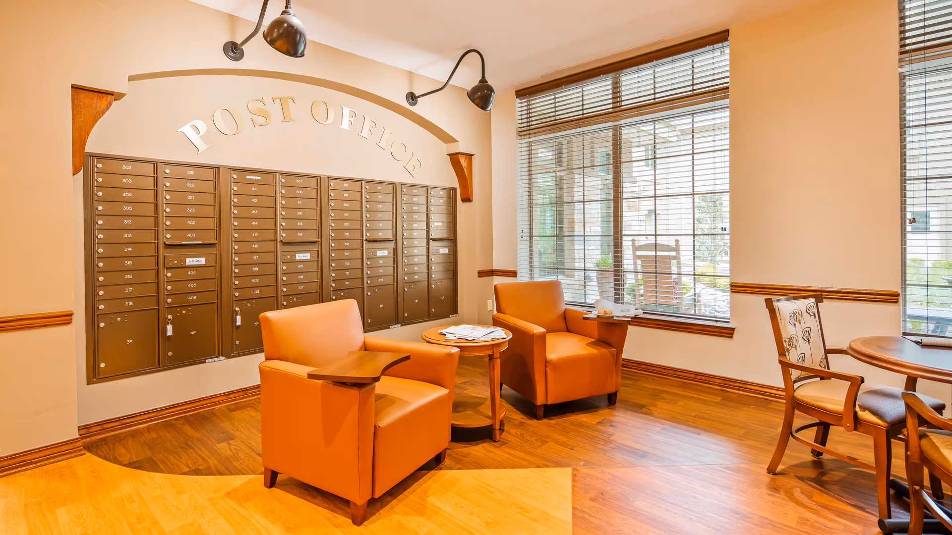 Interior view of a post office area within a facility, featuring a wall of mailboxes labeled 'POST OFFICE'. Two orange armchairs with attached wooden trays are positioned around a small round table with newspapers on it. Large windows with blinds allow natural light to fill the room, and there is a wooden chair and table set near the windows.