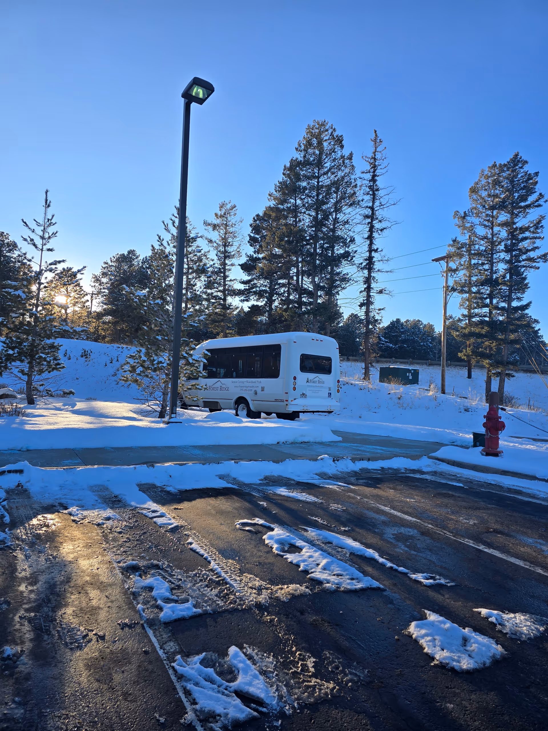 Snow-covered parking lot with a parked white senior-living shuttle bus, lamppost, fire hydrant, and pine trees under a clear blue sky.