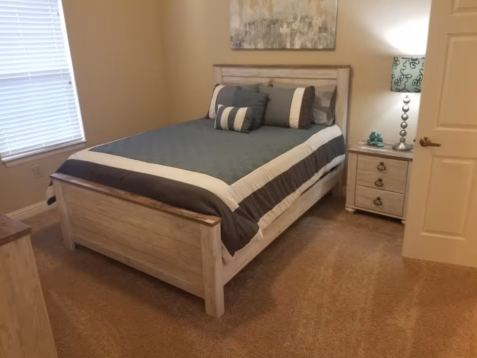 A bedroom with a wooden bed frame and headboard, a neatly made bed with gray and white bedding, a nightstand with a lamp and decorative items, a window with blinds, and a door slightly ajar.