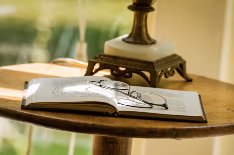 A close-up of a wooden round table with an open book and a pair of eyeglasses resting on it, with a decorative lamp in the background and soft natural light coming through a window.