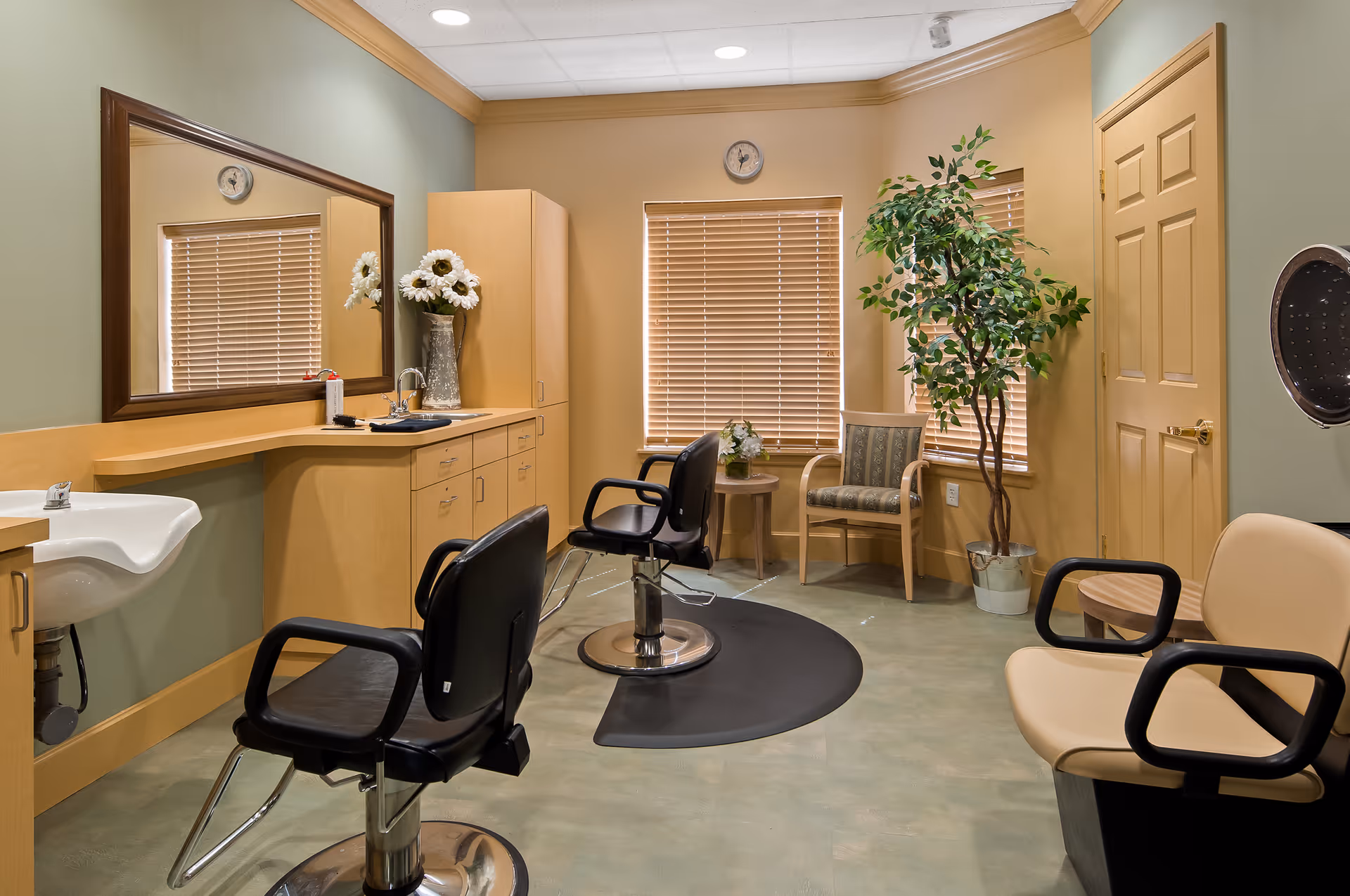 Interior of a salon room in a senior living facility with two black salon chairs in front of a large mirror, a sink, wooden cabinets, a potted plant, and additional seating with beige and patterned chairs near windows with blinds.