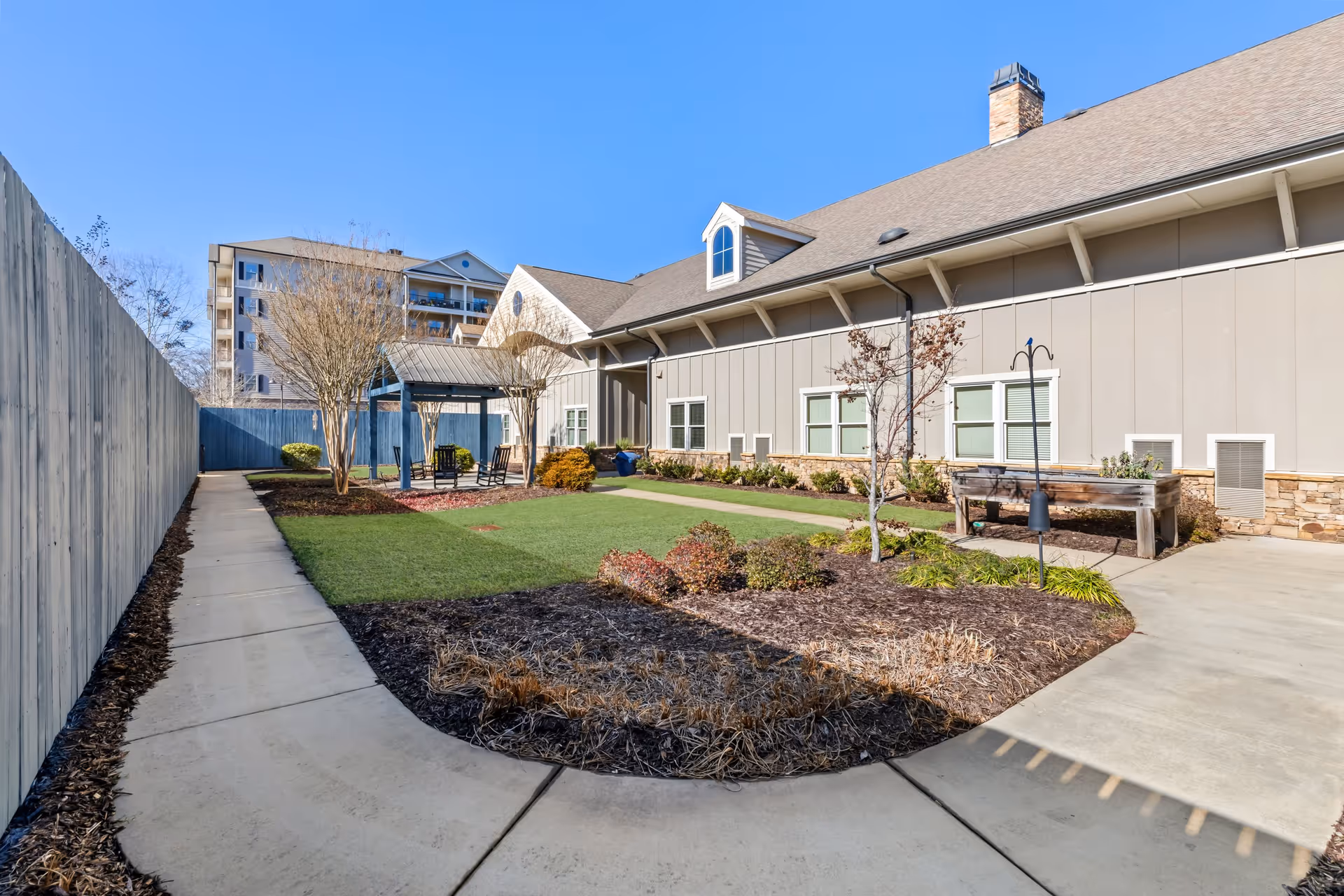Outdoor courtyard area at Arbor Terrace Acworth featuring a paved walkway, landscaped garden beds with shrubs and small trees, a covered seating area with chairs, and a building with beige siding and stone accents under a clear blue sky.