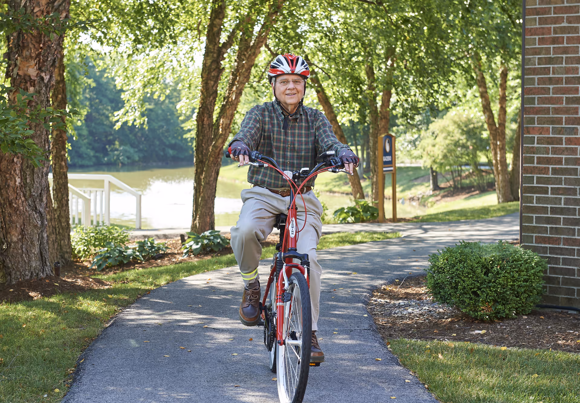 An elderly man wearing a red and white helmet and plaid shirt is riding a red bicycle on a paved path surrounded by trees and greenery near a body of water. There is a brick building and a small bush to the right side of the path.