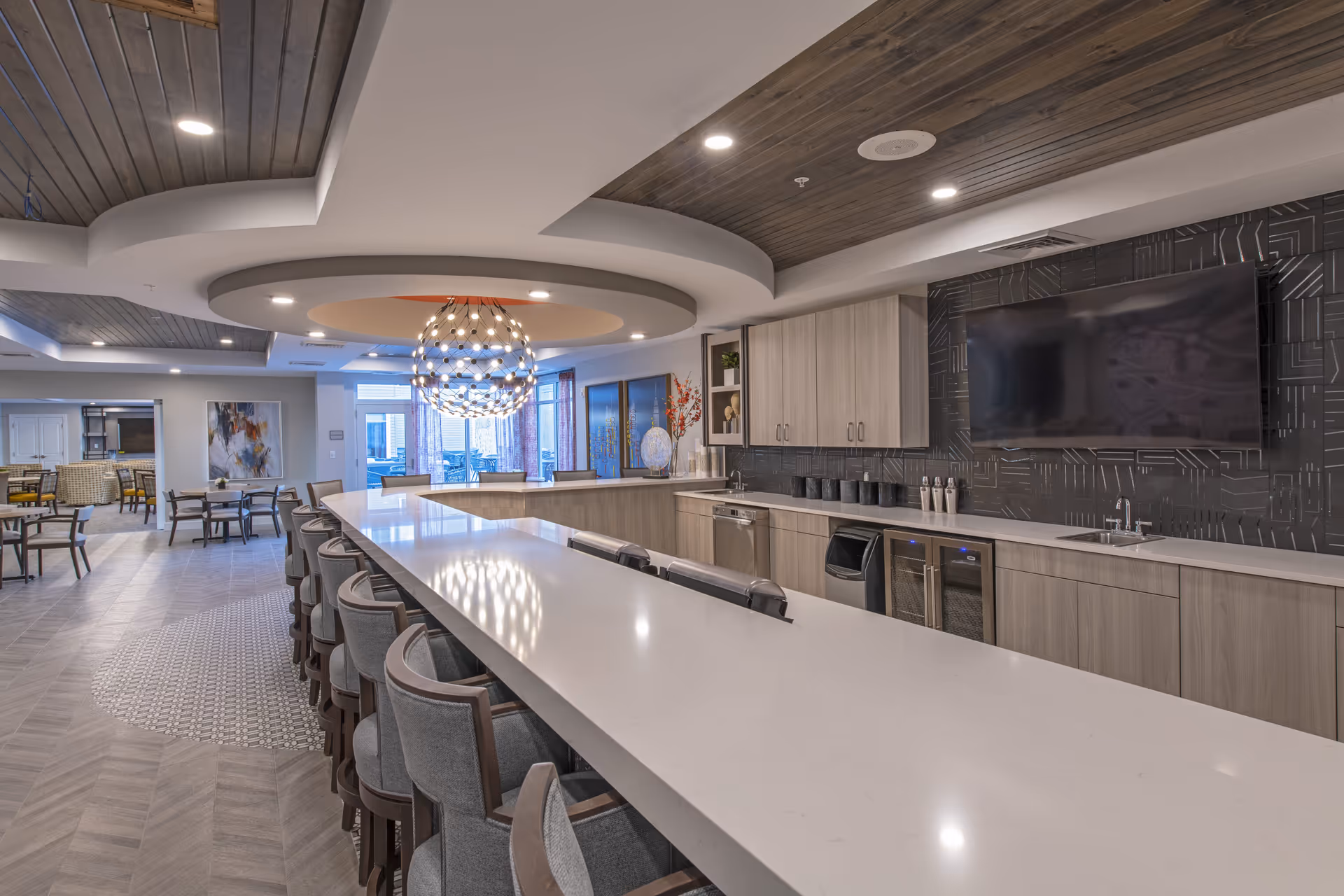 Modern communal kitchen and dining area with a long white countertop surrounded by gray cushioned chairs, wooden cabinetry, a large flat-screen TV mounted on a dark patterned wall, and a decorative spherical chandelier hanging from a recessed ceiling. In the background, there are additional tables and chairs near large windows with curtains.