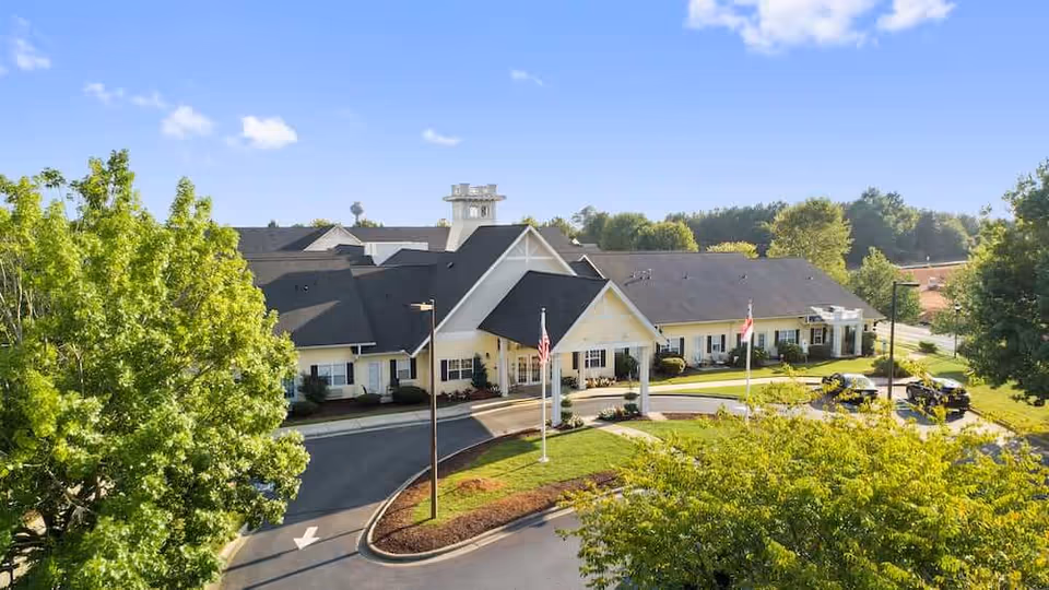 Front entrance of a single-story senior living facility with a covered porte-cochere, circular driveway, flagpoles, and surrounding trees under a blue sky.