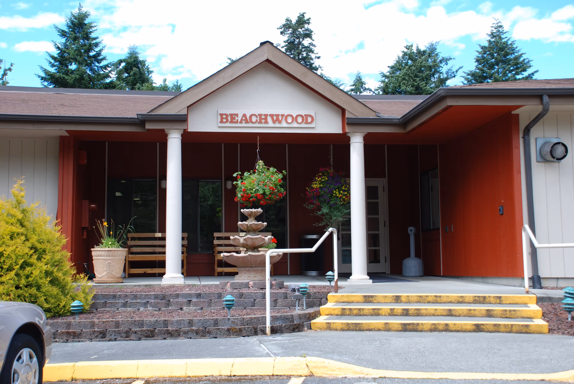 Entrance to a building with a sign reading 'BEACHWOOD' above the doorway. The entrance has white columns, a three-tiered stone fountain, hanging flower baskets, benches, and steps with yellow safety markings leading up to the door. There are trees and a partly cloudy sky in the background.