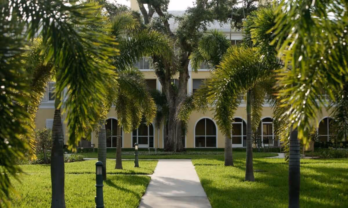 A paved walkway lined with palm trees leads to a yellow building with arched windows and a balcony, surrounded by well-maintained green grass and landscaping.