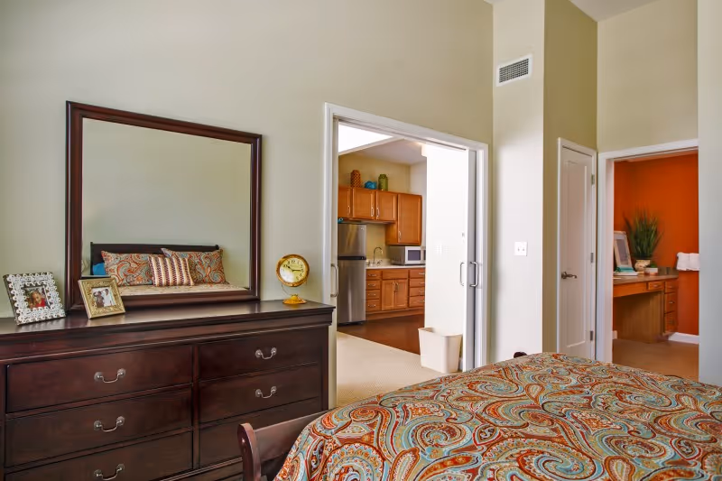 A bedroom with a dark wooden dresser topped with a large mirror, two framed photos, and a clock. The bed has a colorful paisley-patterned bedspread and matching pillows. Through an open doorway, a kitchen with wooden cabinets, a stainless steel refrigerator, and a microwave is visible. Another doorway leads to a room with an orange accent wall and a desk with a plant and framed picture.