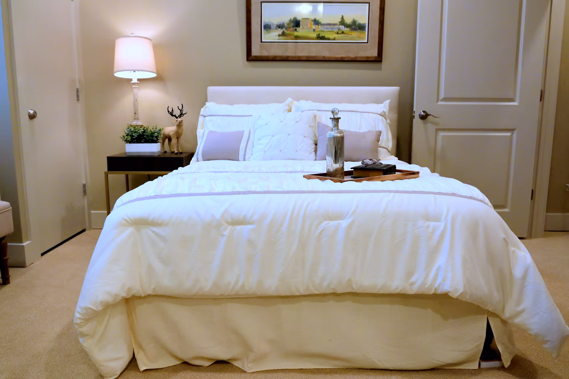 A neatly made bed with white bedding and two light purple pillows in a bedroom. A wooden tray with a glass bottle and some books is placed on the bed. To the left of the bed is a nightstand with a lamp, a small plant, and a decorative deer figurine. There are two closed doors on either side of the bed, and a framed picture hangs on the wall above the headboard.