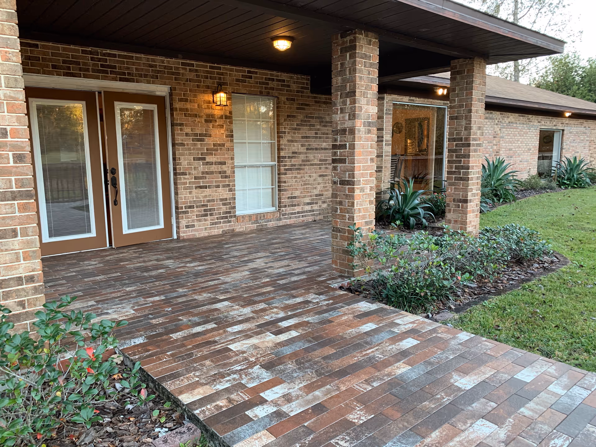 Covered brick patio area with brick columns and a tiled floor outside a building. There are glass double doors and windows with blinds, outdoor light fixtures, and landscaped plants along the edge of the patio and lawn.