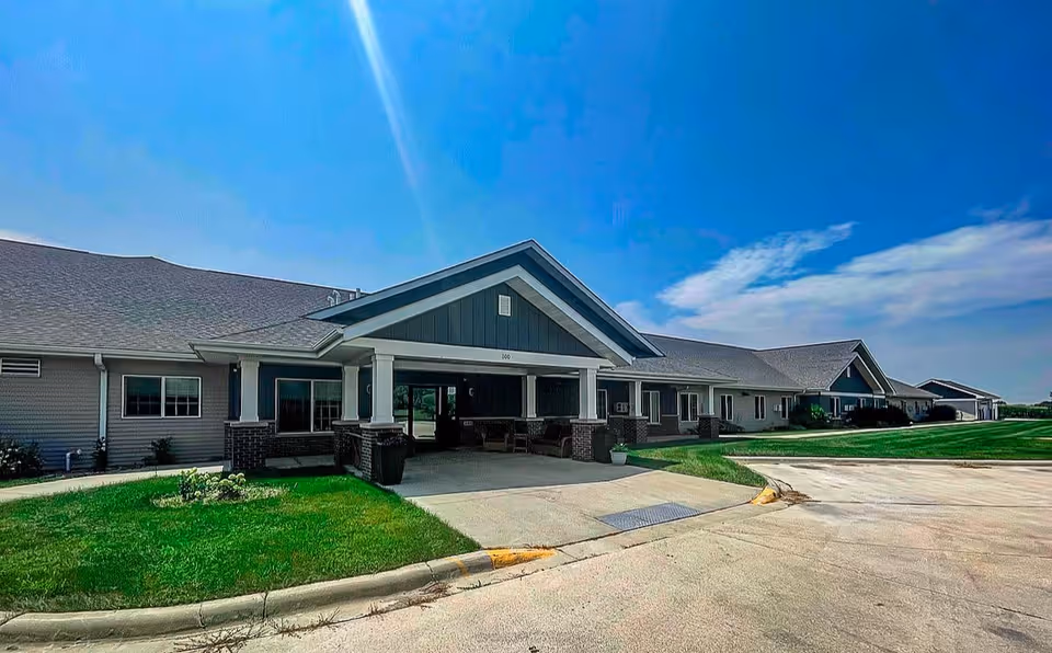 Exterior view of a single-story senior living facility building with a covered entrance, green lawn, and a clear blue sky with some clouds.