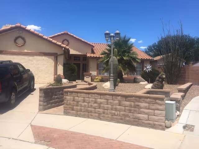 Front exterior of a single-story stucco home with a tiled roof, low brick front wall, desert landscaping and a lamppost beside a driveway with a parked SUV.