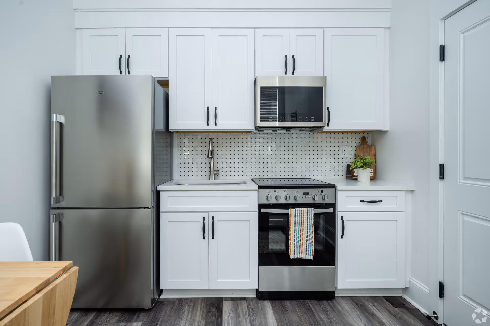 Modern kitchen with white cabinets, a stainless steel refrigerator, a built-in microwave above a stove with a colorful striped towel hanging on the oven handle, a sink with a faucet, and a small plant on the countertop. The floor is wood-style laminate and the backsplash has a white tile pattern with small black squares.