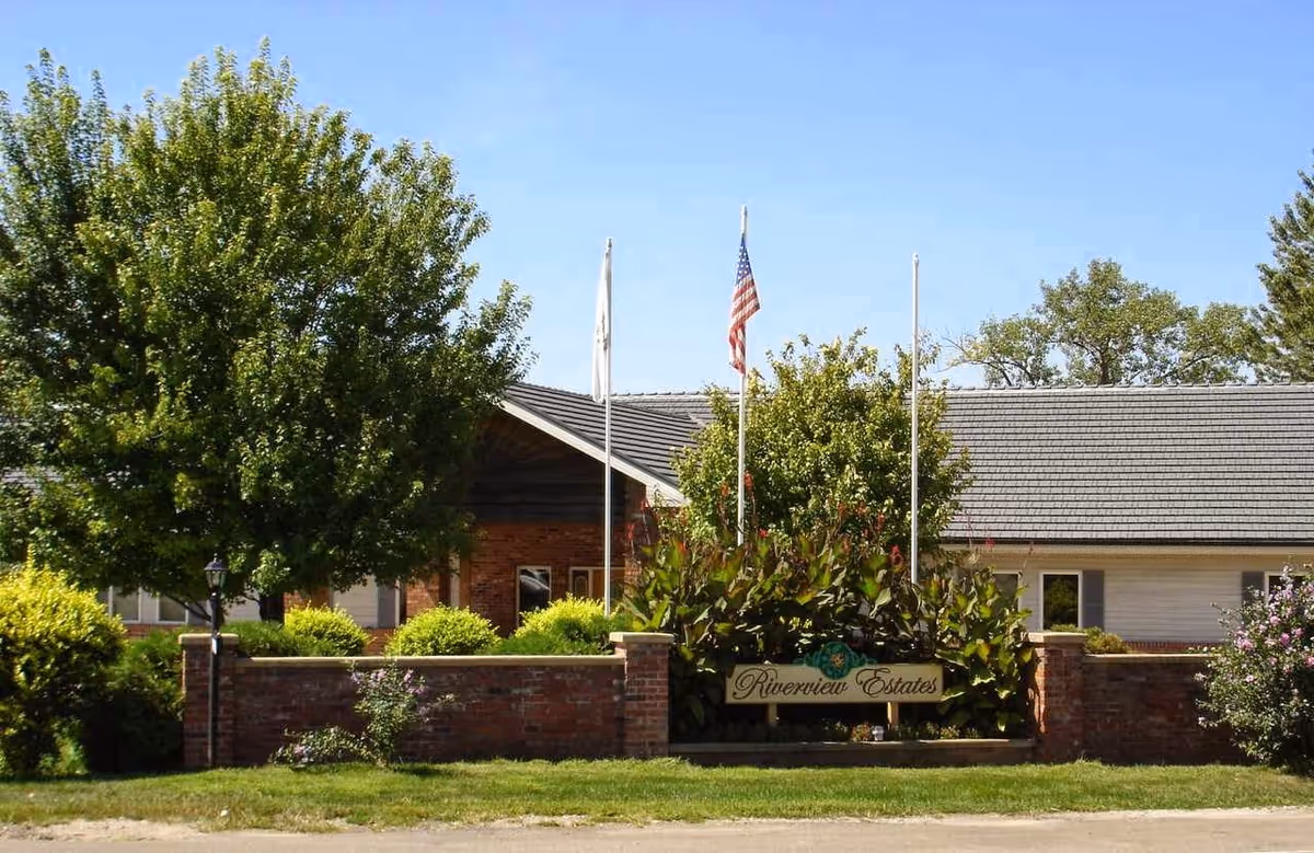 Exterior view of a senior living facility named Riverview Estates with a brick wall and sign in front, surrounded by green trees and bushes under a clear blue sky.