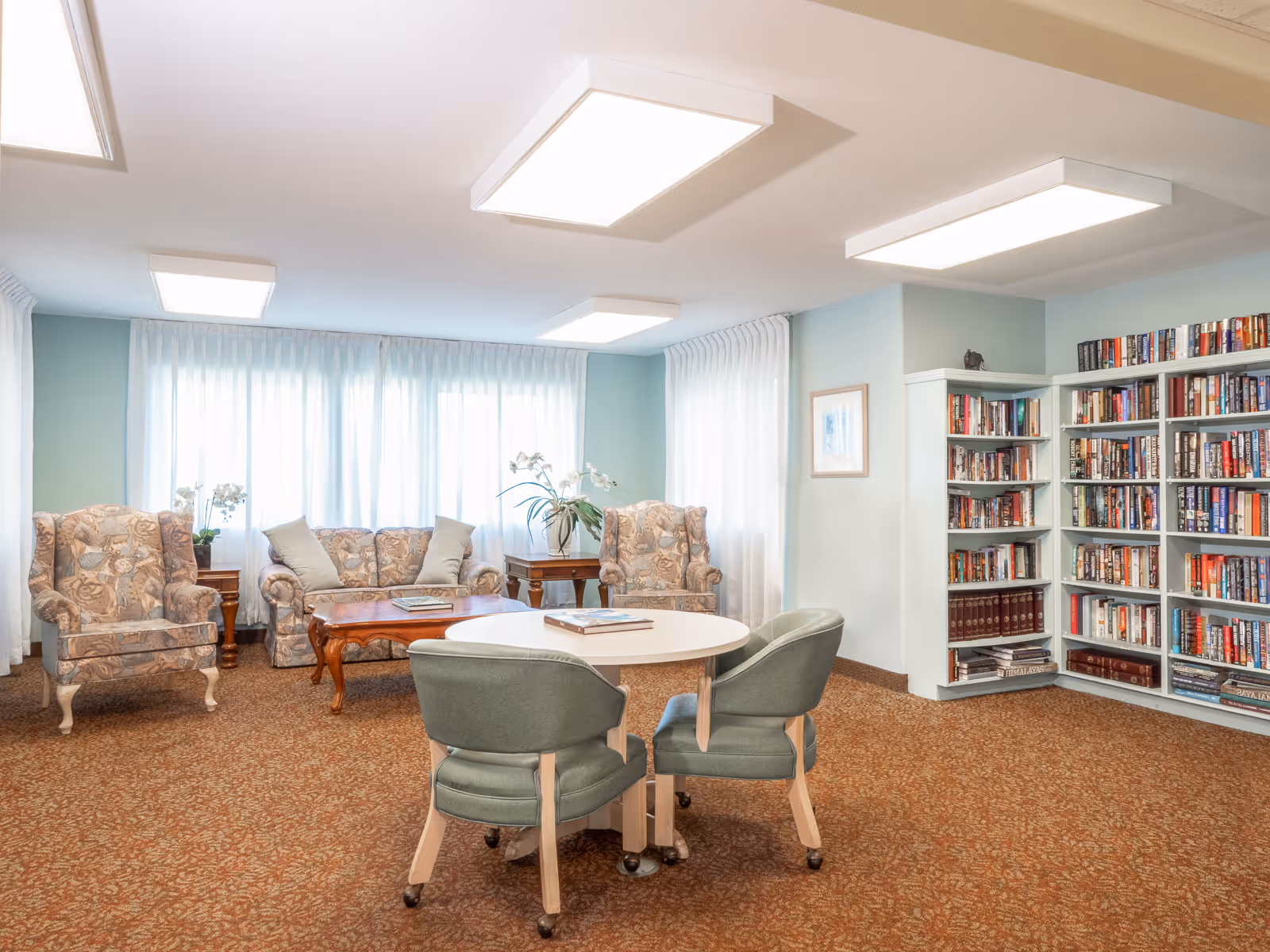 A bright lounge with upholstered armchairs, a sofa, a round table with chairs, and bookshelves along the wall.