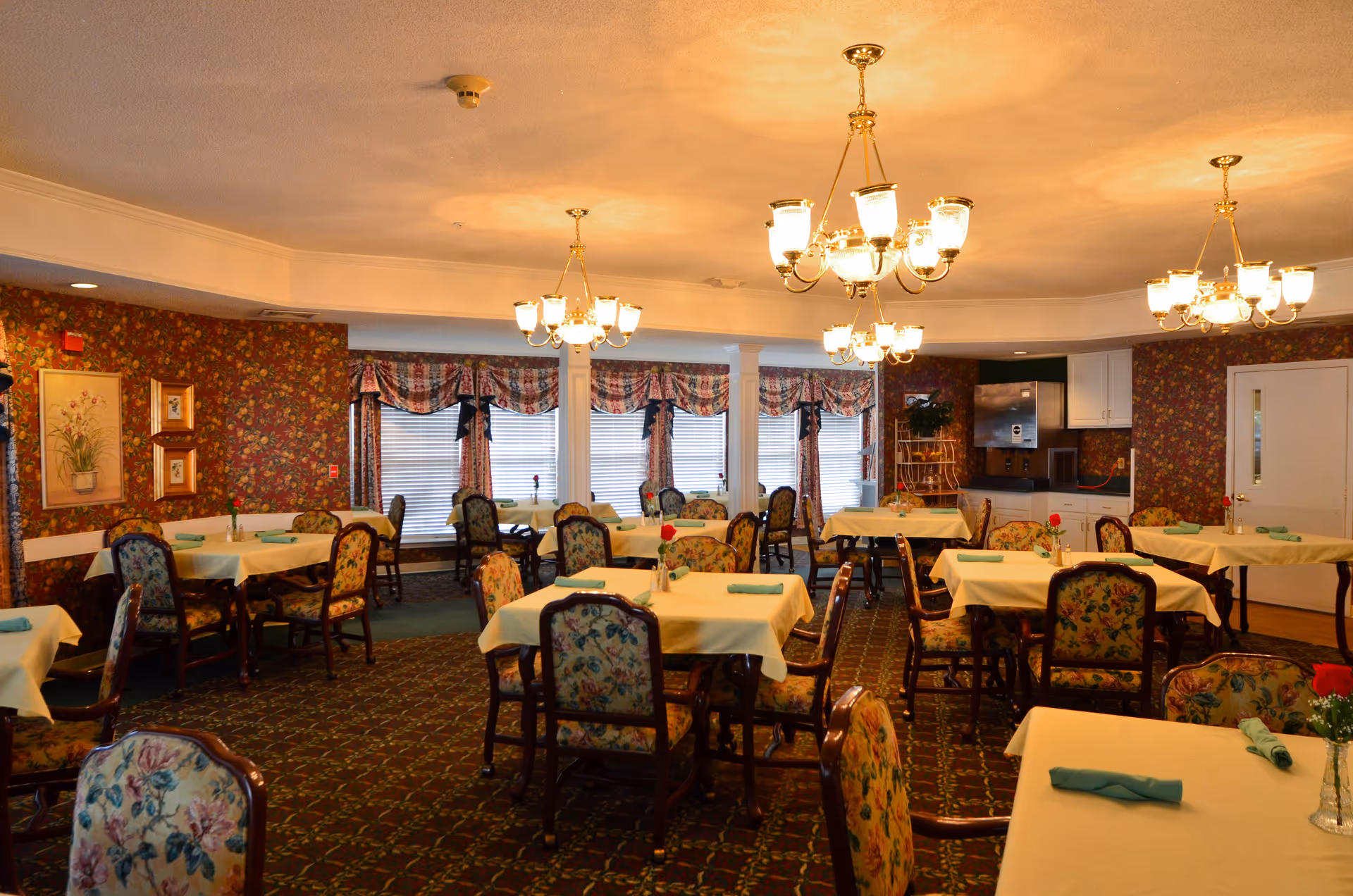 A dining room with multiple tables covered in yellow tablecloths, each set with a rolled green napkin and a small vase with a red flower. The chairs have floral upholstery. The room has floral wallpaper, patterned carpet, and several chandeliers hanging from the ceiling. Large windows with floral curtains line one wall.