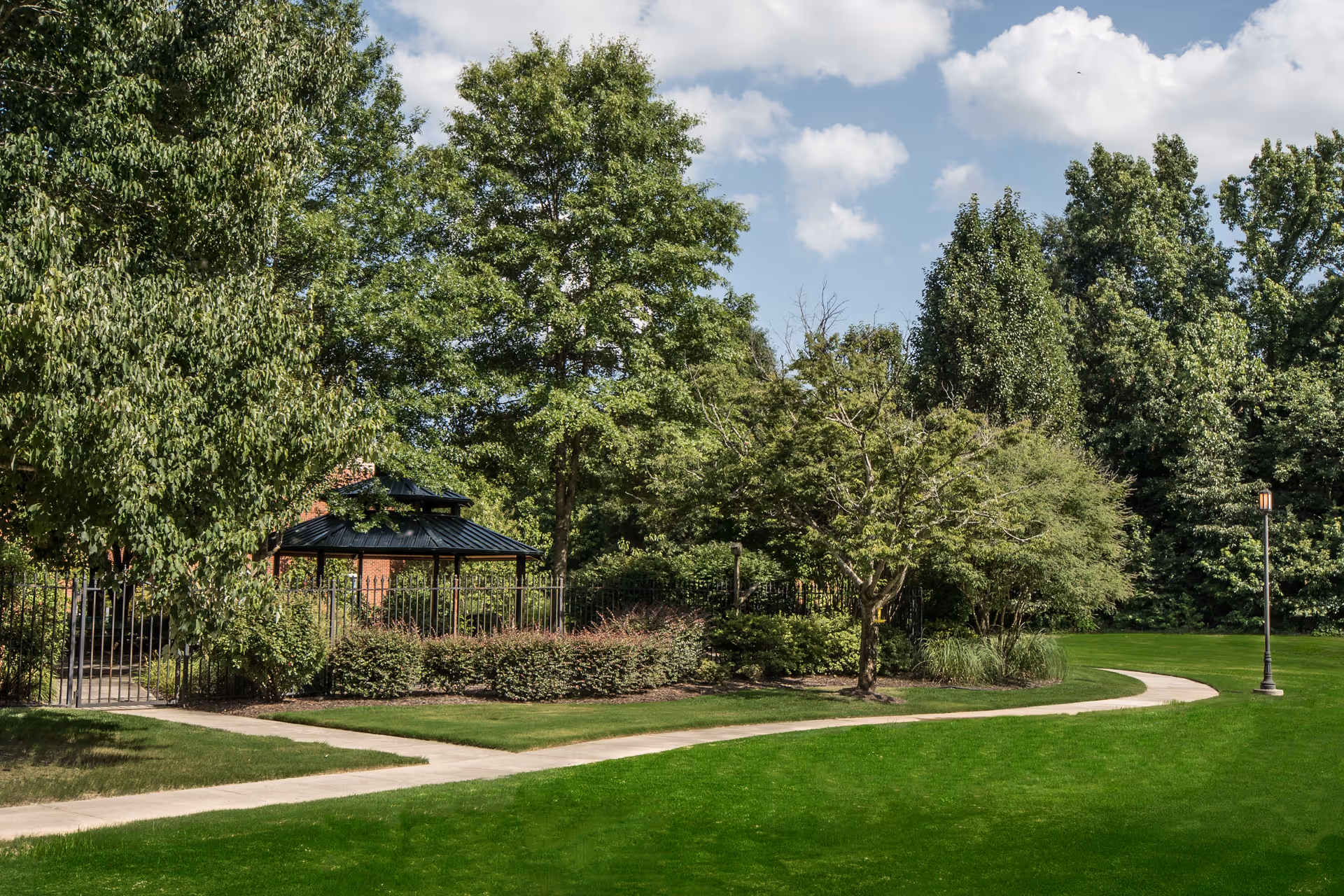 A well-maintained outdoor garden area with a paved walking path, green grass, various trees, bushes, and a black metal gazebo surrounded by a fence under a partly cloudy sky.