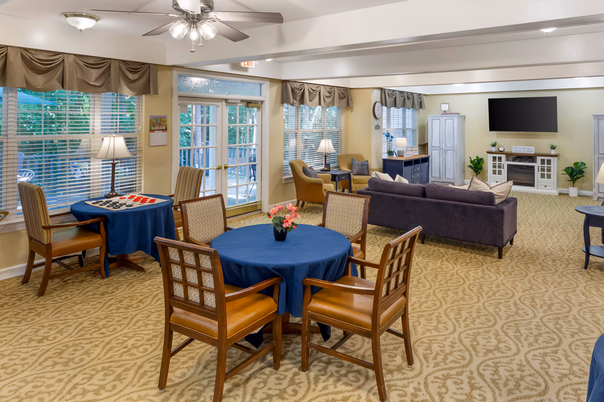 A spacious and well-lit common area with patterned carpet flooring, featuring a round table with a blue tablecloth and four wooden chairs in the foreground. To the left, there is a smaller table with a blue tablecloth and two striped chairs, set up for a board game. The background shows a seating area with a dark purple couch, two beige armchairs, side tables with lamps, and a large flat-screen TV mounted on the wall above a white entertainment unit. Large windows with blinds and brown valances let in natural light, and glass doors lead to an outdoor area with greenery visible outside.