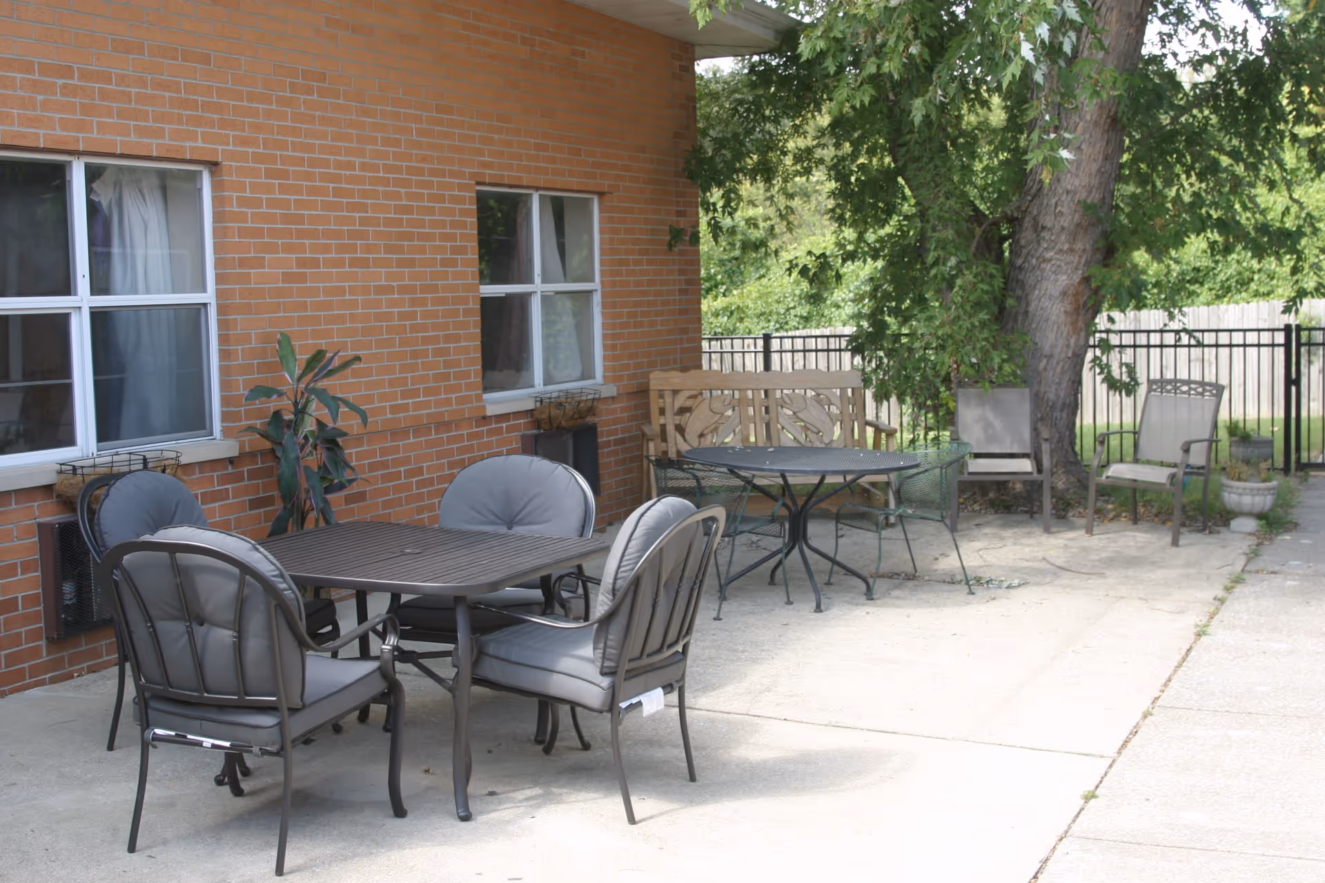 Outdoor patio area with multiple seating arrangements including a table with four cushioned chairs, a round table with metal chairs, a wooden bench, and two additional chairs near a large tree. The area is adjacent to a brick building with two windows and surrounded by greenery and a black metal fence.