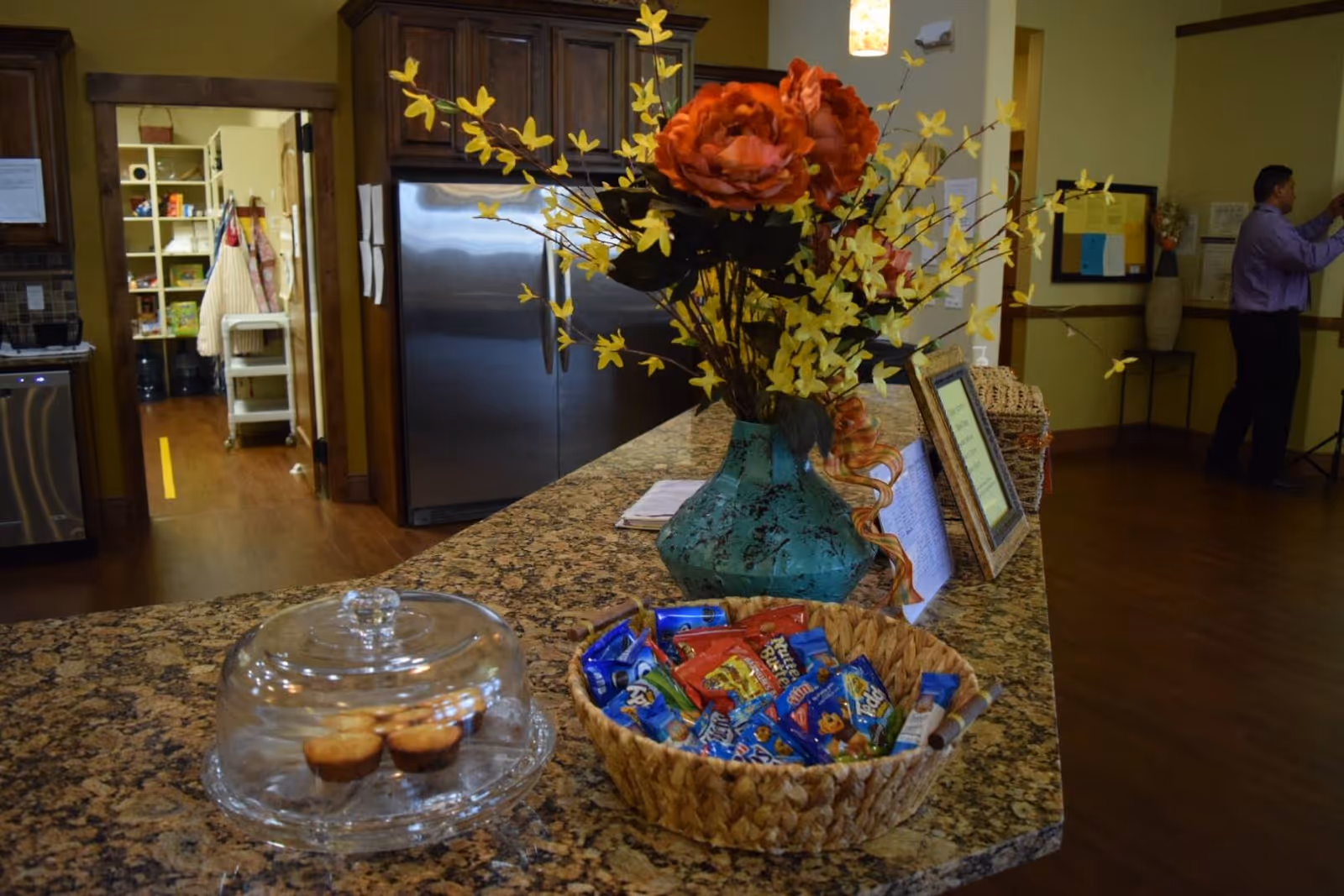 A kitchen or dining area countertop with a basket of assorted snacks and a glass dome covering three muffins. Behind the counter is a large stainless steel refrigerator and wooden cabinets. A decorative vase with orange and yellow flowers is placed on the counter. In the background, a man in a purple shirt is standing near a wall with bulletin boards and a small table with a vase.