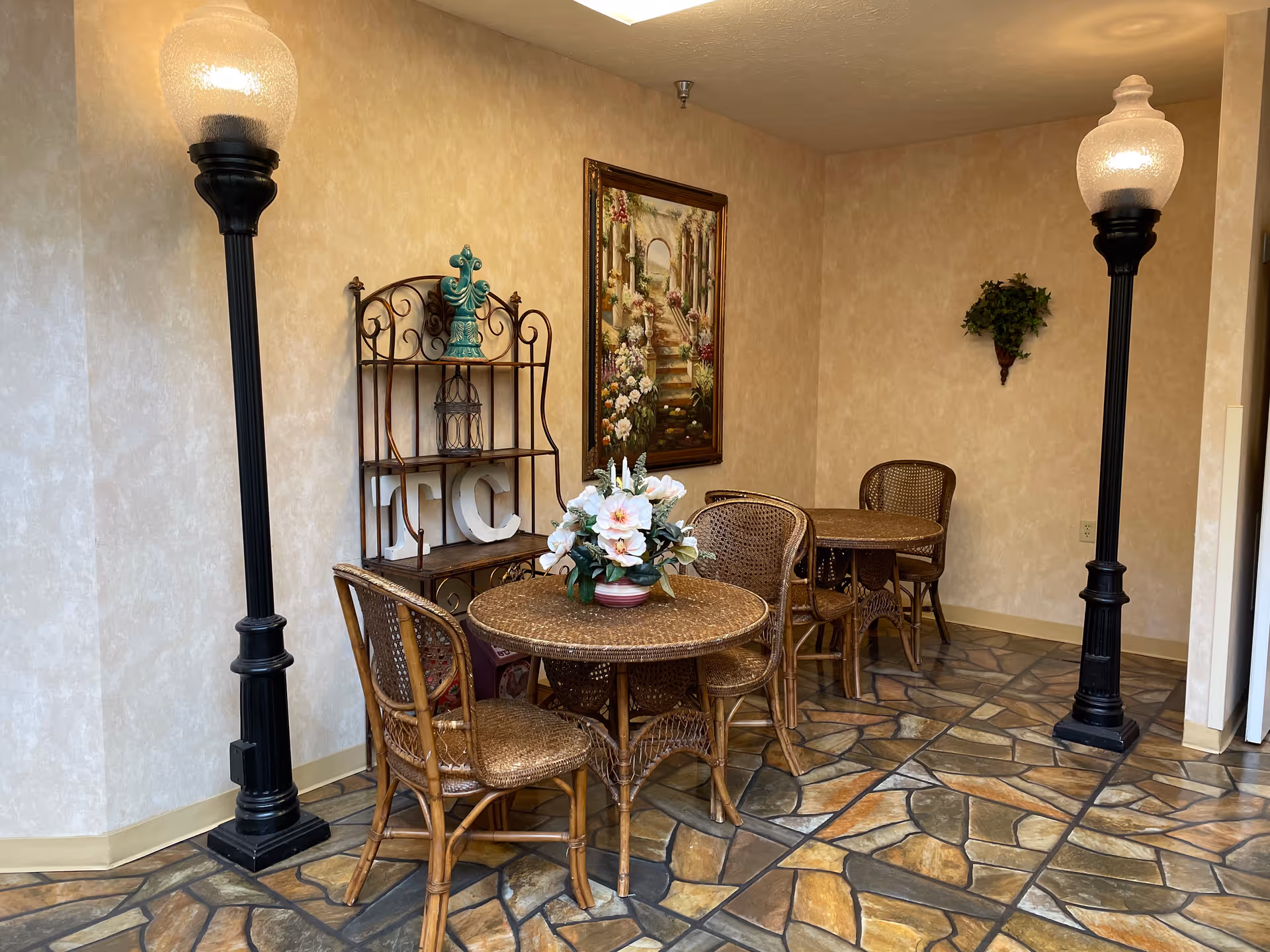 Small seating area with wicker tables and chairs, a floral centerpiece, decorative shelving and wall art between two tall lamp posts on a tiled floor.