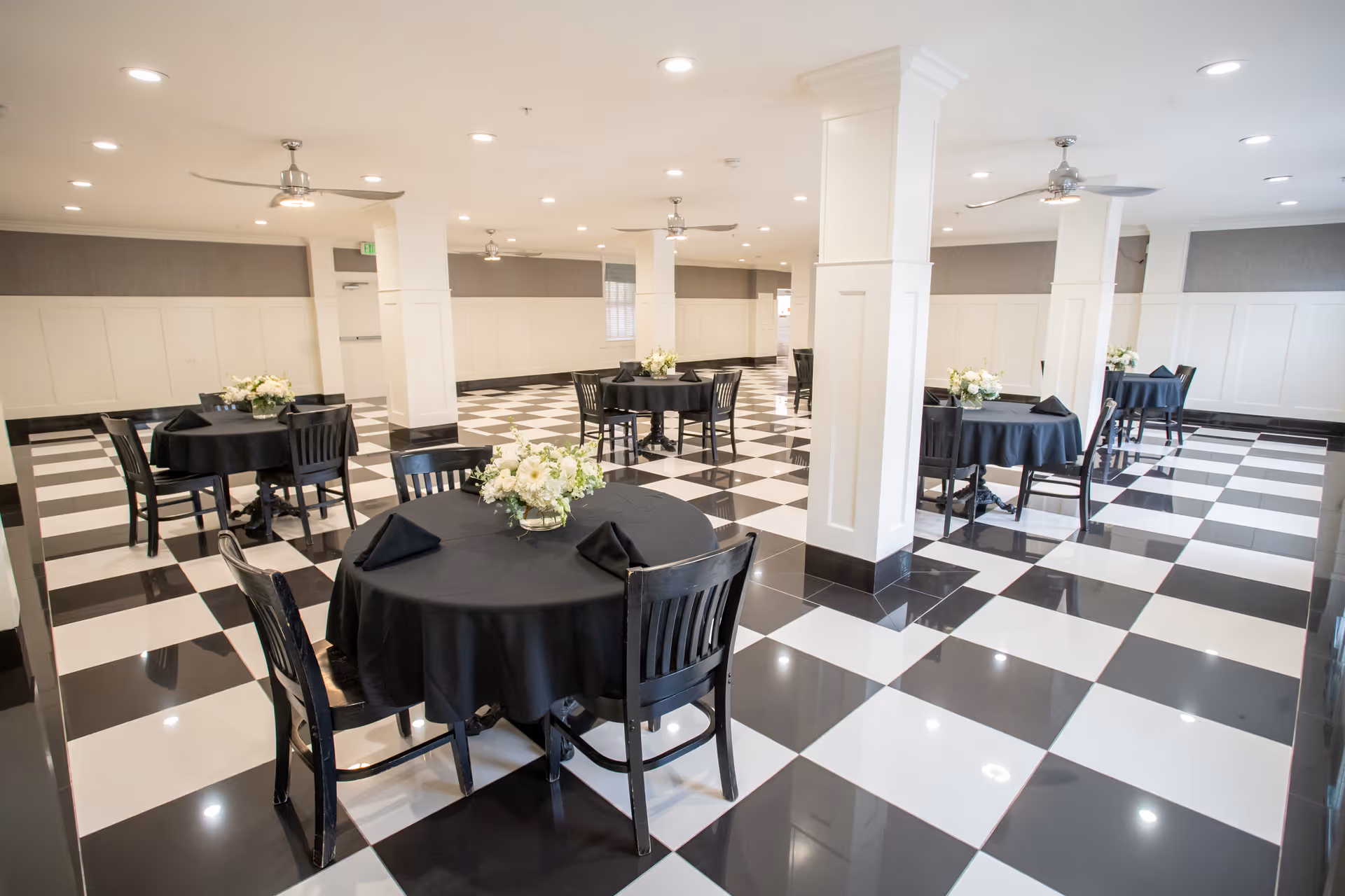A spacious dining room with black and white checkered tile flooring, round tables covered with black tablecloths, black chairs, and floral centerpieces on each table. The room has white paneled walls, several white columns, ceiling fans, and recessed lighting.