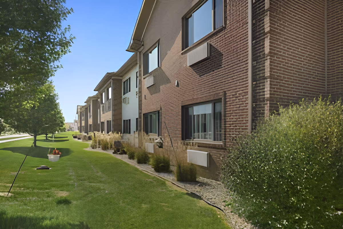 Exterior view of a multi-unit residential building with brick walls and several windows. There is a well-maintained grassy area with bushes and trees alongside the building under a clear blue sky.