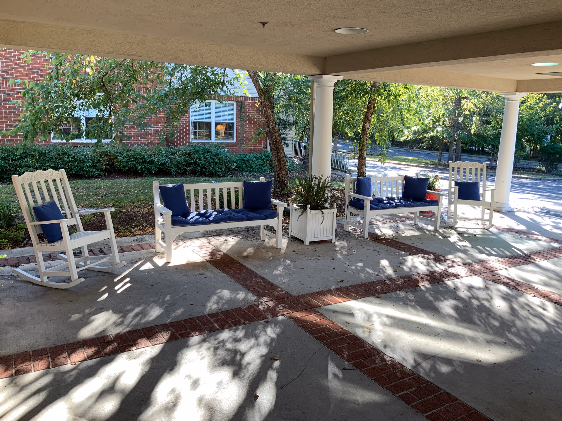 Covered outdoor seating area with white rocking chairs and benches featuring blue cushions, surrounded by trees and greenery with a brick building in the background.