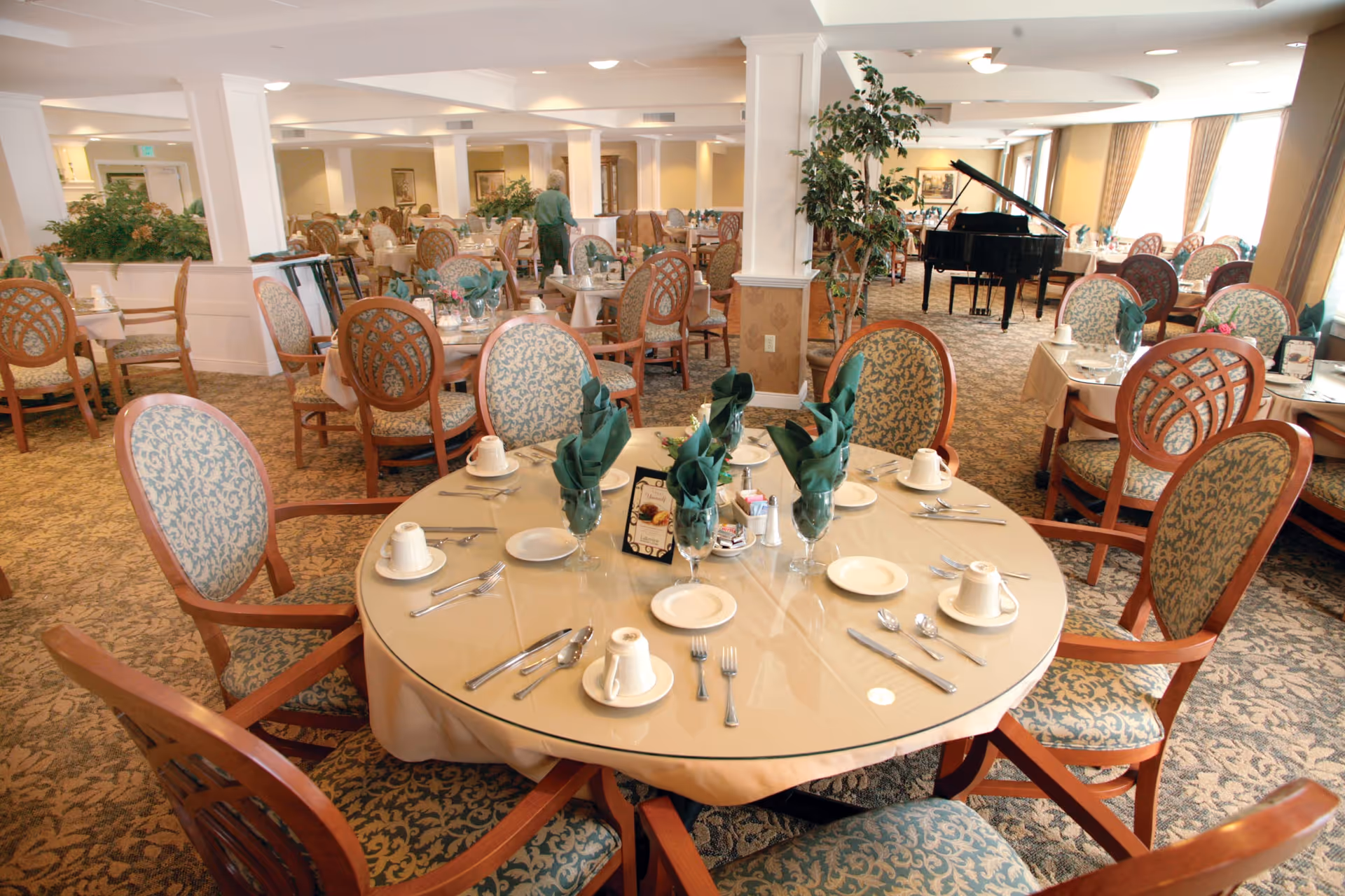 A spacious dining room in a senior living facility with multiple round tables set with plates, cups, silverware, and green folded napkins. The room features patterned carpet, upholstered wooden chairs, large windows with curtains, and a black grand piano in the background. A person is seen walking in the distance.