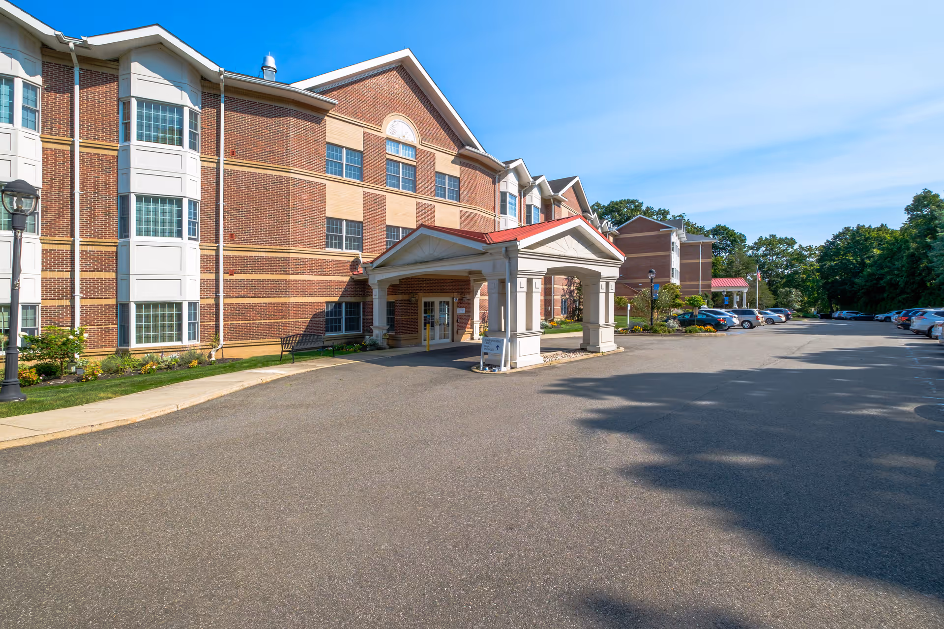Front entrance of a multi-story red brick senior living building with a covered porte-cochère and a parking lot.