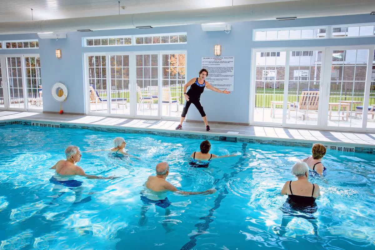 A group of elderly people participating in a water exercise class in an indoor swimming pool. An instructor stands at the poolside demonstrating movements. Large windows and glass doors let in natural light and show an outdoor patio area with chairs.