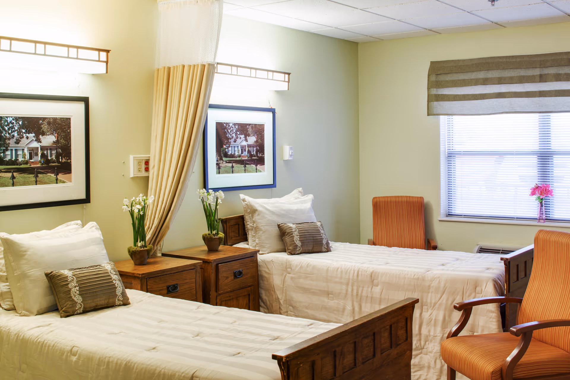 A well-lit bedroom with two single beds featuring wooden frames and white bedding. Each bed has a decorative pillow and is separated by a wooden nightstand with a small potted plant. The room has light green walls, two framed pictures above the beds, a window with blinds and a striped valance, and two orange upholstered chairs near the window.
