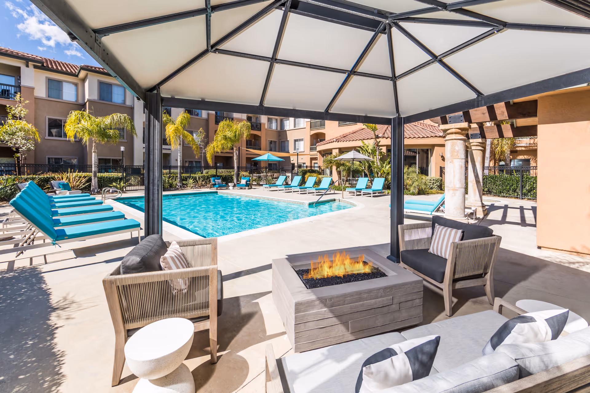 Outdoor pool area at a senior living facility with turquoise lounge chairs arranged around the pool. In the foreground, there is a shaded seating area with cushioned chairs and a fire pit. The building with balconies and palm trees surrounds the pool area under a clear blue sky.