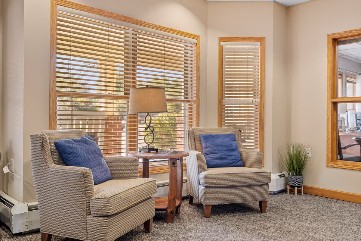 A cozy seating area in a senior living community featuring two cushioned armchairs with blue pillows, a wooden side table with a decorative lamp, large windows with wooden blinds, beige walls, and a small potted plant in the corner.