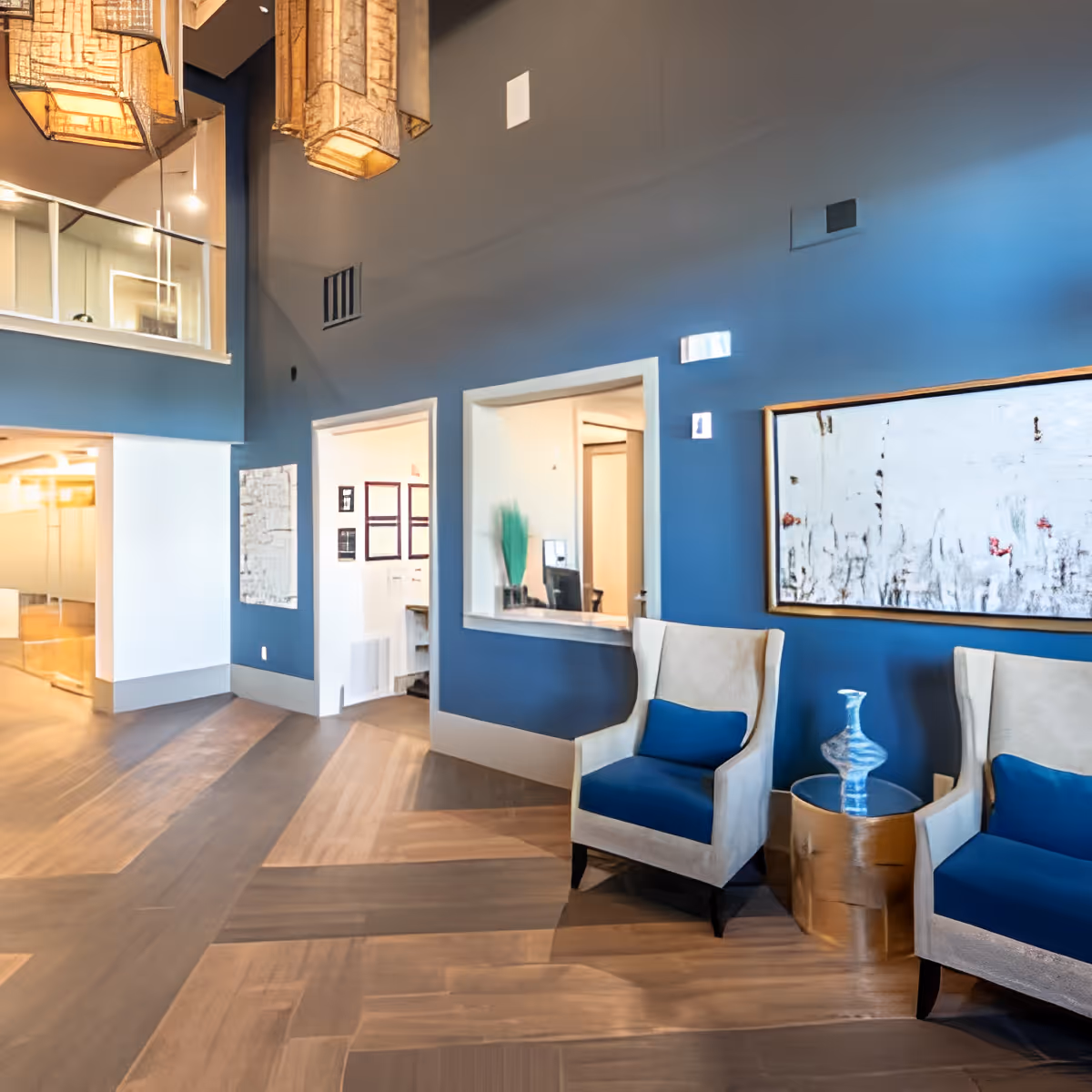 Interior view of a senior living facility lobby area with two beige armchairs featuring blue cushions, a small round wooden table with a decorative glass vase, blue walls, a large abstract painting, and modern hanging light fixtures. There is a reception window and an open doorway leading to another room.
