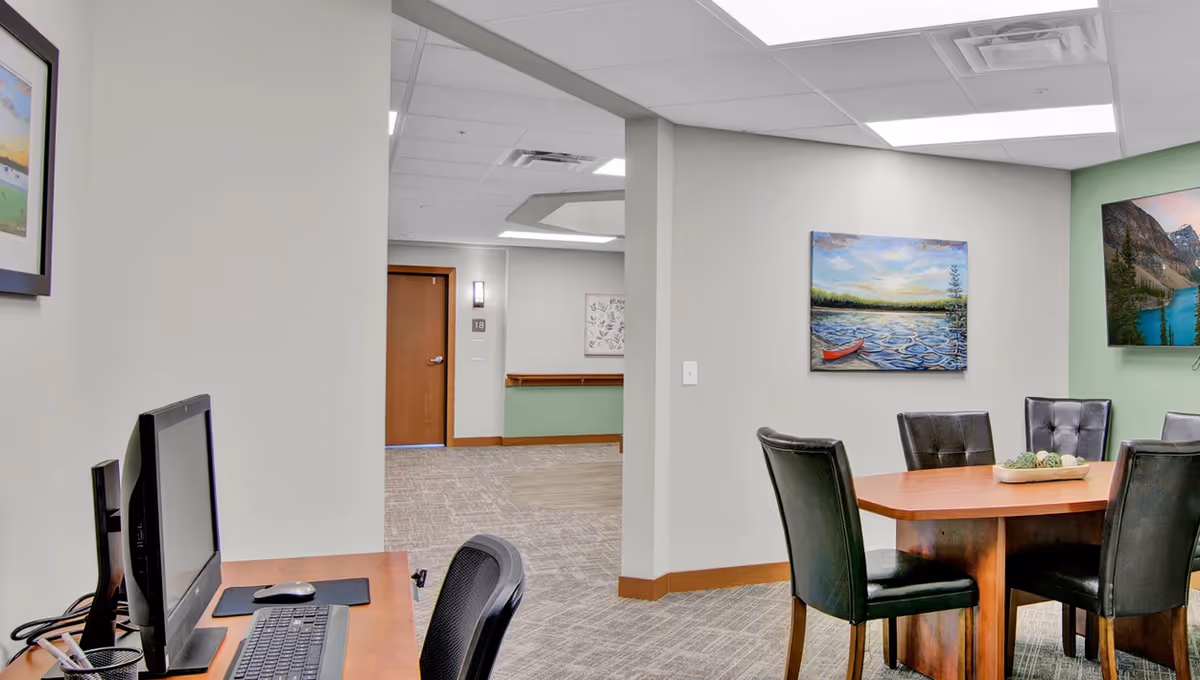 Interior view of a senior living facility showing a workspace with a computer and keyboard on a wooden desk to the left, and a wooden table with six black leather chairs on the right. The walls are decorated with landscape paintings, and there is a hallway with a wooden door labeled 18 in the background.