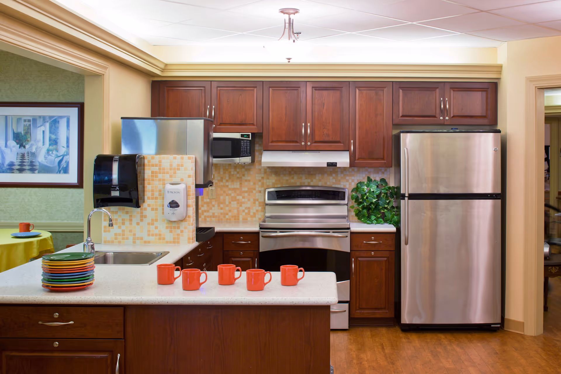 A kitchen area with wooden cabinets, a stainless steel refrigerator, oven, and microwave. There is a countertop with a sink, a stack of colorful plates, and five orange mugs. The backsplash has a mosaic tile design, and there is a green plant on the counter near the refrigerator. A paper towel dispenser and hand sanitizer dispenser are mounted on the wall. In the background, a dining area with a yellow tablecloth and framed artwork is partially visible.