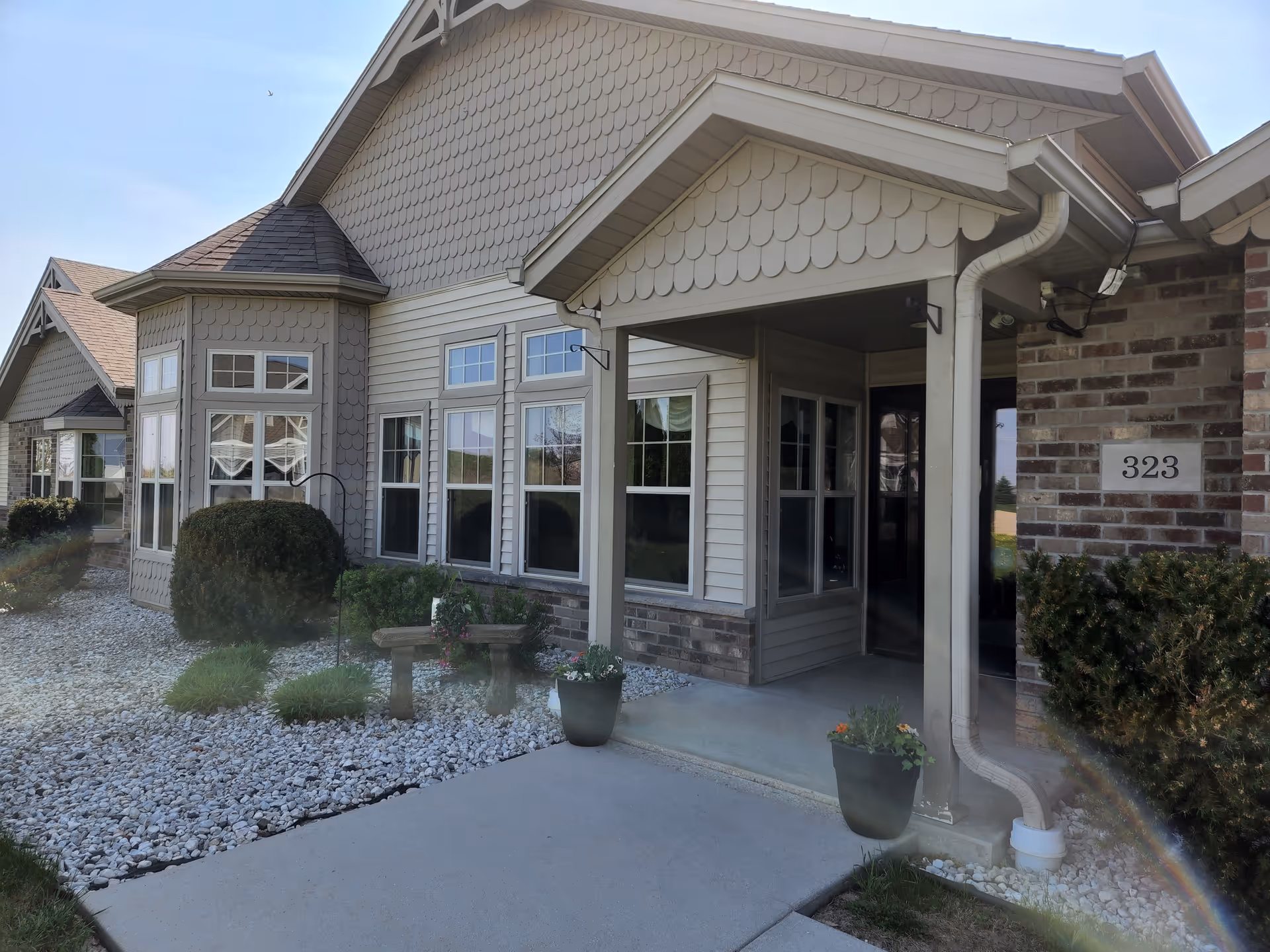 Exterior view of a residential-style building with beige siding, brick accents, and multiple windows. There is a covered entrance with two potted plants on either side of the walkway, bushes, and white decorative rocks in the landscaping. The building number 323 is visible on the brick wall near the entrance.
