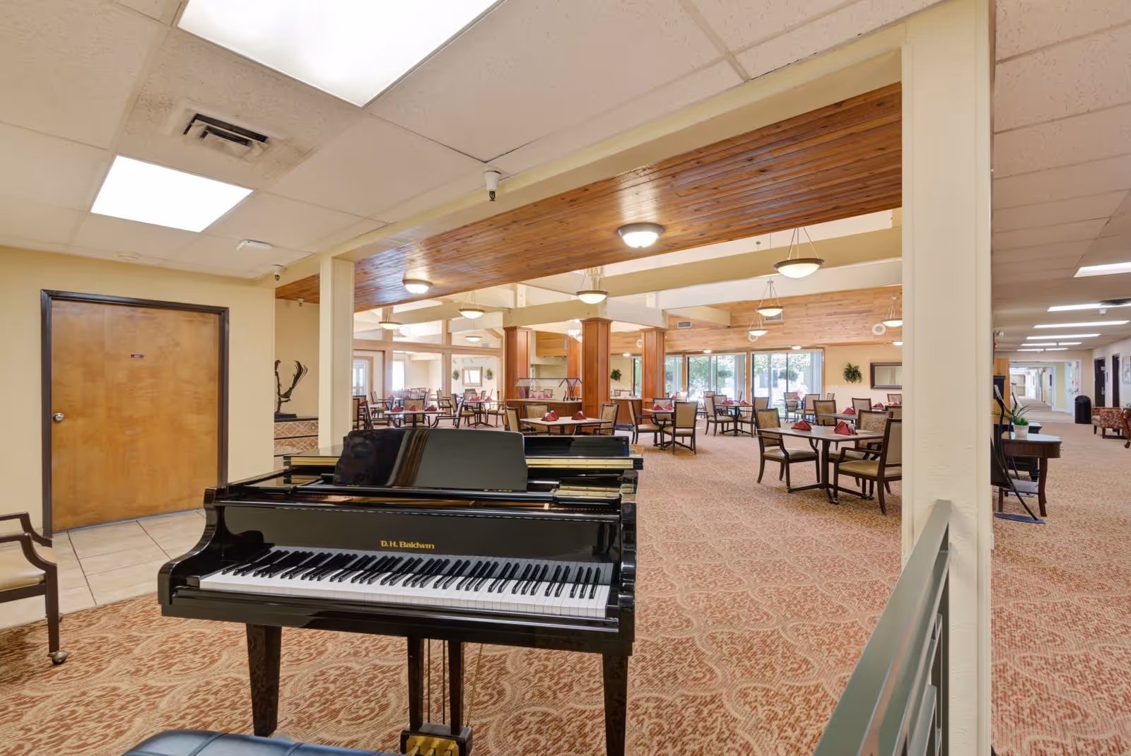 Interior view of a retirement community common area featuring a black grand piano in the foreground, multiple tables and chairs arranged for dining or socializing, beige walls, carpeted floor with a patterned design, and ceiling lights with wooden accents.