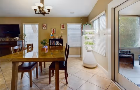 Interior view of a dining area with a wooden table and chairs, a chandelier overhead, a TV on a stand, a bookshelf with decorative items, a vertical indoor garden planter with plants, and a glass door leading to an outdoor patio.
