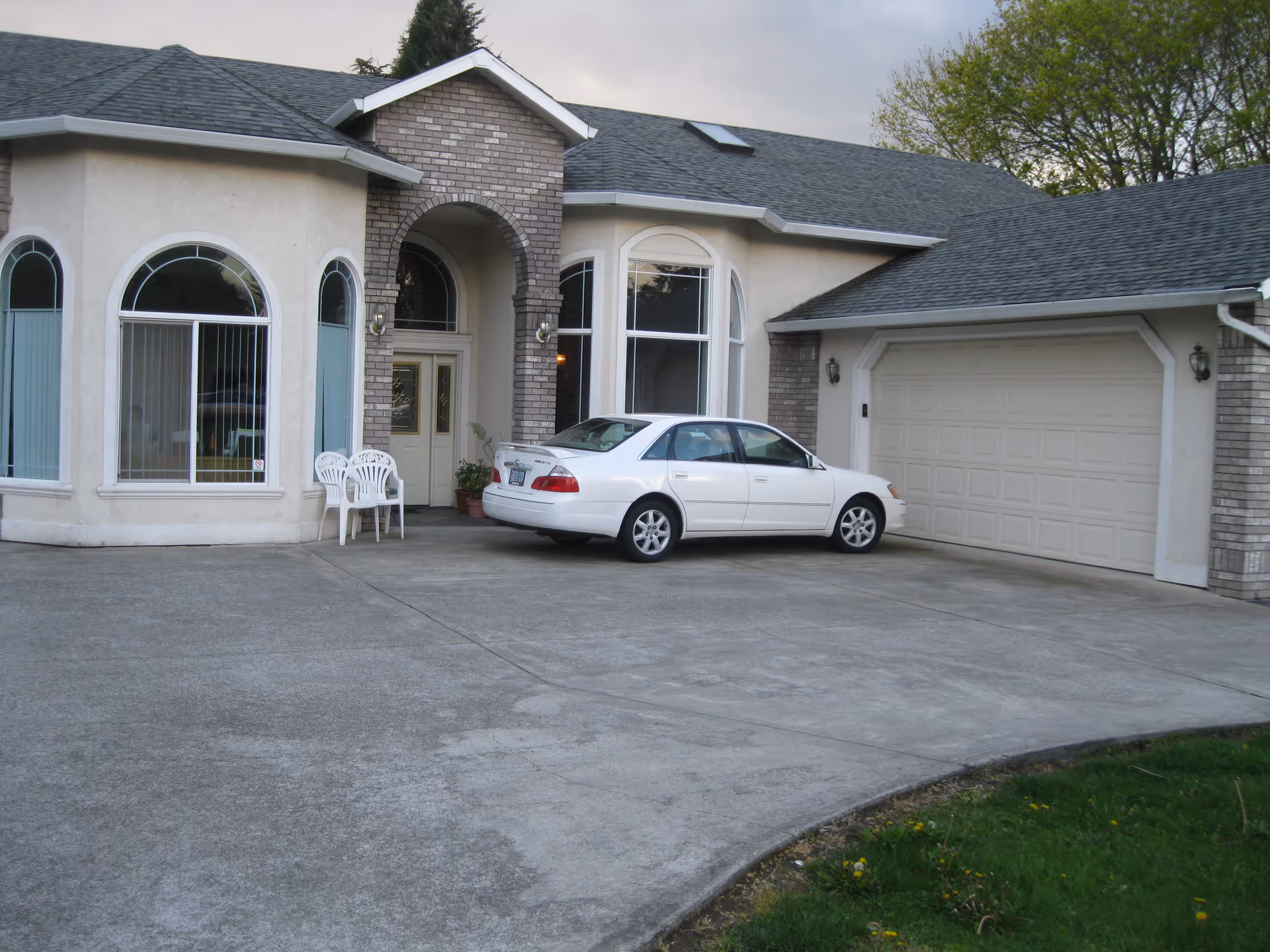 Exterior view of a single-story residential building with a large driveway, a white car parked in front of a closed garage door, and two white plastic chairs near the entrance. The building has arched windows and a brick accent around the front door.